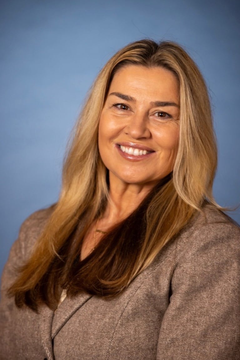 Professional headshot of a smiling woman with long golden-brown hair wearing a taupe blazer against a blue background
